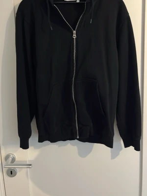 Svart hoodie med dragkedja från Weekday - Säljer en svart hoodie från Weekday i storlek S Hoodien har dragkedja framtill, två stora fickor och justerbar huva med snören. Tillverkad i mjukt bomullsmaterial och har ribbade muddar vid ärmslut och nederkant. Perfekt för en chill och avslappnad stil.