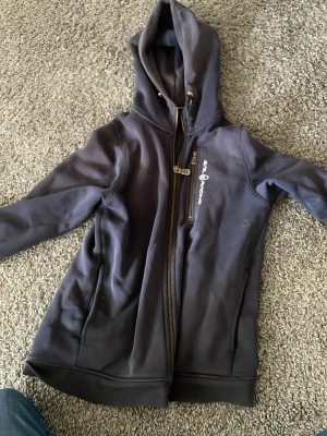Mörkblå sail racing zip hoodie - Snygg mörkgrå hoodie med huva och dragkedja framtill. Har en bröstficka med dragkedja och tryckt logga på vänster sida. Tillverkad i mjukt material, perfekt för chill eller utehäng. Långa ärmar och normal passform. Jag är 170 på bilden och enligt mig är den för liten! Skulle säga bäst är vid 158 och 164cm passar den som bäst och är använd👍🔥