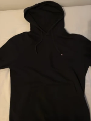 Svart hoodie från Tommy Hilfiger - Snygg svart hoodie från Tommy Hilfiger med liten logga på bröstet och klassisk huva med dragsko. Tillverkad i mjukt bomullsmaterial som känns skönt mot huden. Perfekt för en avslappnad och stilren look.