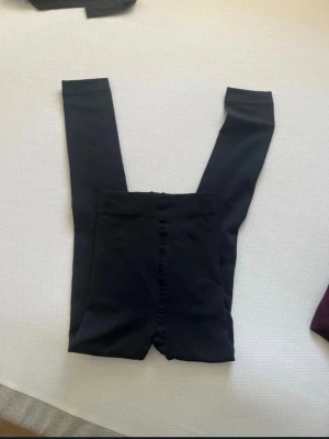 Svarta leggings med hög midja - Säljer ett par svarta leggings med hög midja och knappar framtill. Leggingen är i stretchigt material och har en slät, matt finish. Perfekta att bära under kjol eller klänning för extra komfort.