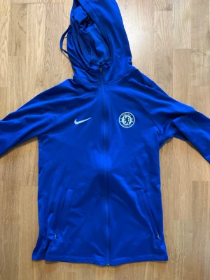Blå Chelsea Nike hoodie - Snygg blå hoodie med dragkedja från Nike och Chelsea FC-logga på bröstet. Tröjan har huva med dragsko, två fickor med dragkedja och är tillverkad i ett sportigt, mjukt material. Perfekt för träning eller när du vill visa ditt stöd för Chelsea.