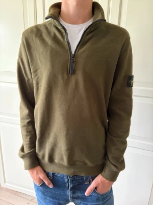 Olivgrön halfzip från Stone Island - Snygg olivgrön tröja från Stone Island med halv dragkedja och hög krage. Tröjan har lång ärm och den klassiska Stone Island-loggan på vänster ärm. Tillverkad i mjuk bomull, perfekt för lager på lager och streetwear. Tröjan är i storlek M. Hör av er vid frågor och priset är inte hugget i sten!
