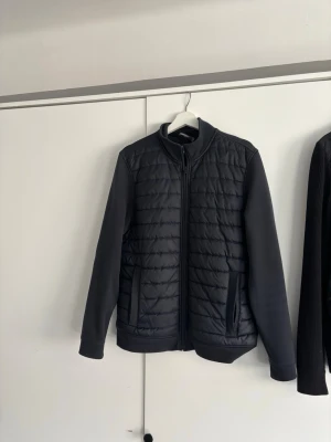 Mörkblå pufferjacka med dragkedja - Snygg mörkblå pufferjacka med quiltad framsida och släta ärmar. Jackan har hög krage, två sidofickor med dragkedja och stängs med en hel dragkedja framtill. Perfekt för kalla dagar och ger en stilren look.