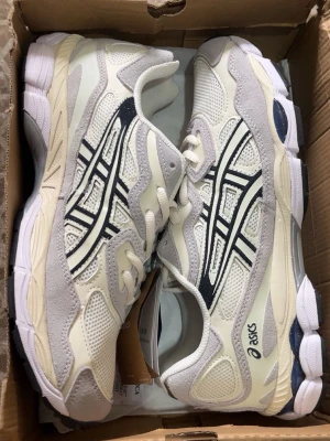 Ascis  - Snygga vita sneakers från Asics med detaljer i beige och grå mocka. Skorna har meshpaneler för extra andningsförmåga och klassiska svarta Asics-loggor på sidorna. Sulan är vit med inslag av grått och blått. Perfekta för en sportig och trendig look.