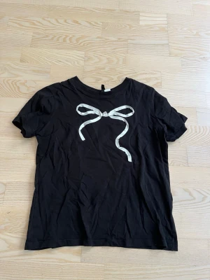 Svart t-shirt med rosettdetalj - Säljer en svart t-shirt i bomull med korta ärmar och en vit rosettdetalj framtill. Enkel och stilren design som passar perfekt till jeans eller kjol. Skön passform och mjukt material.