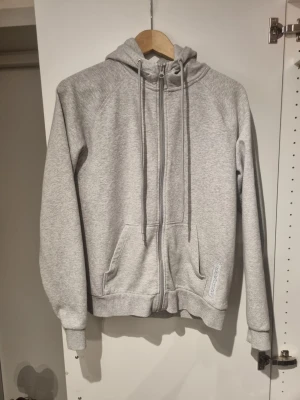 Grå zip hoodie Peak Performance - Snygg grå zip hoodie från Peak Performance med huva och dragsnören. Klassisk modell med fickor framtill och ribbade muddar vid ärmar och nederkant. Perfekt för en avslappnad och sportig stil. Tillverkad i mjukt sweatshirtmaterial.