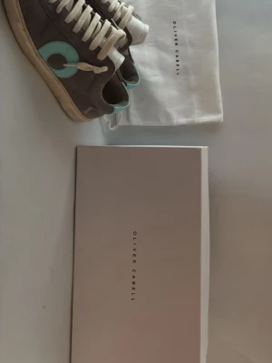 Oliver Cabell sneakers grå/turkos - Säljer ett par sneakers från Oliver Cabell i grå mocka med turkosa detaljer och vita skosnören. Skorna har en unik cirkeldesign på sidan och turkos hälflik med logga. Medföljer originalpåse och låda. De ända defekterna är ena hälen och toe boxen. Skorna tvättas innan de skickas och priset kan diskuteras