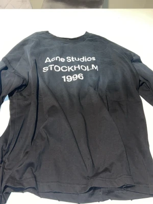 Acne studios tröja - Svart långärmad från Acne Studios med vit text 'STOCKHOLM 1996' på bröstet. Klassisk rund hals och avslappnad passform. Perfekt för dig som gillar stilrena och trendiga plagg.