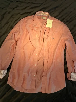 Rosa skjorta från Gant med knappar  - Säljer en rosa skjorta från Gant med klassisk button-down krage och vita knappar. Skjortan har lång ärm och randiga detaljer på insidan av manschetterna. Perfekt för en stilren look. (originalpris på denna skjorta är 1400kr, kolla sista bilden för att se😊)