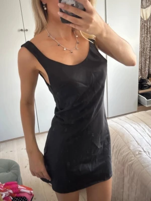 Svart slip dress - Snygg svart kortklänning med breda axelband och rundad urringning. Klänningen är figurnära och har en enkel, stilren design som passar perfekt till många tillfällen. Materialet ser ut att vara mjukt och följsamt.