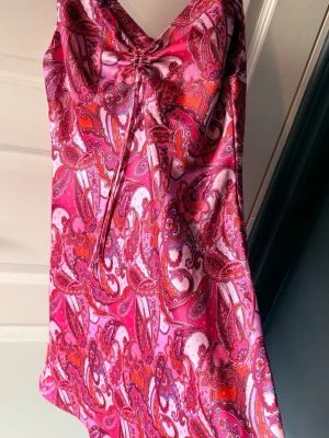 Rosa paisleymönstrad kort klänning - klänning i siden material med paisleymönster i rosa, rött och lila toner. Klänningen har en rosett vid bysten. Är från bik bok i storlek S, knappt använd 