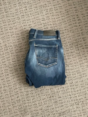 Replay Anbass 30/32 - Snygga blå slim fit jeans från Replay med klassisk femficksdesign och slitna detaljer på låren. Jeansen har kontrastsömmar och en skinnpatch med logga bak i midjan. Perfekta för dig som gillar en modern och avslappnad stil.