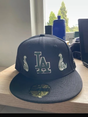 Svart LA New Era 59FIFTY keps - Säljer en svart snapback keps från New Era, modell 59FIFTY, med LA-logga och två paisleymönstrade detaljer framtill. Kepsen har rak skärm och broderad New Era-logga på sidan. Perfekt för dig som gillar streetwear och vill sticka ut.