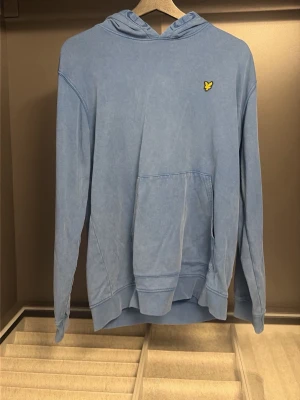 Ljusblå lyle scott hoodie  - Snygg ljusblå hoodie med huva och känguruficka framtill. På bröstet sitter en liten gul logga som detalj. Hoodien har långa ärmar och avslappnad passform, perfekt för en chill look. Tillverkad i mjukt bomullsmaterial.