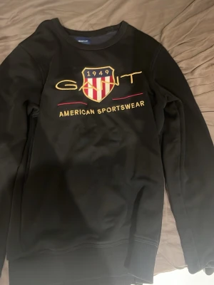 Svart sweatshirt från Gant - Svart sweatshirt från Gant med broderad logga och sköld på bröstet. Texten 'American Sportswear' i gult och rött ger en sportig vibe. Tröjan har rund hals och ribbade muddar. Perfekt för en avslappnad och stilren look.