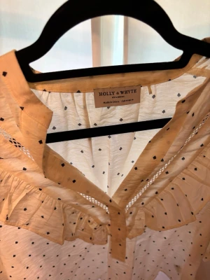 Prickig blus med volang Holly & Whyte - Söt ljusbeige blus från Holly & Whyte med små svarta prickar och bred volang längs ringningen. Blusen har trekvartsärm och knappar framtill. Tillverkad i ett lätt och luftigt tyg, perfekt för en trendig och avslappnad look.