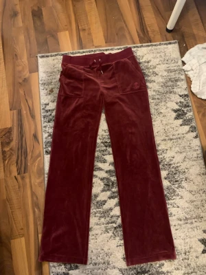 Vinröda velour mjukisbyxor - Snygga vinröda mjukisbyxor i mjuk velour med raka ben och bred resår i midjan. Byxorna har dragsko, stora fickor fram och bakfickor med knappdetaljer. Perfekta för en chill och avslappnad stil.