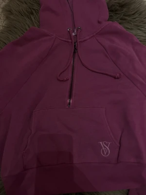 Victoria Secret hoodie - Säljer en mörk rosa victoria secret hoodie med halv dragkedja framtill, huva med dragsko och stor magficka. På fickan finns en broderad VS-logga. Hoodien är gjord i mjukt bomullsmaterial.