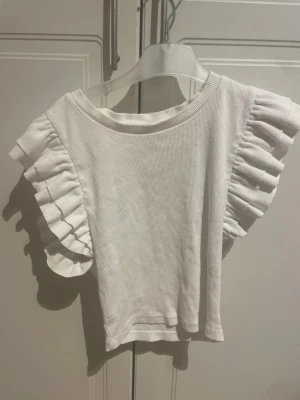 Vit topp med volangärmar från Zara - Supersöt vit topp från Zara med breda volangärmar och ribbad struktur. Perfekt för dig som vill ha en enkel men ändå unik topp till garderoben. Materialet känns mjukt och toppen har en normal passform.