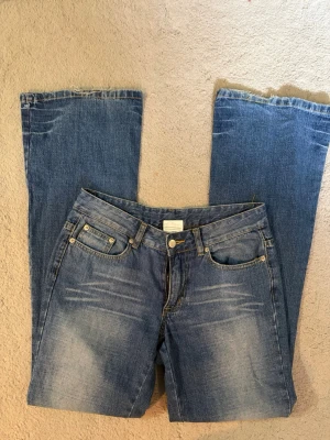 Blå bootcut jeans med slitningar - Säljer ett par klassiska blå bootcut jeans med snygga slitningar framtill och bakfickor med dekorativa sömmar. Jeansen har fem fickor, bälteshällor och stängs med knapp och dragkedja. Perfekta för en avslappnad och trendig look.