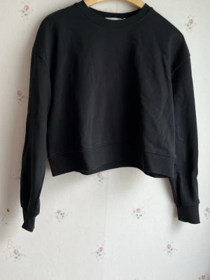 Svart croppad sweatshirt - En stilren svart croppad sweatshirt med rund hals och långa ärmar. Tröjan har ribbade muddar vid ärmslut och nederkant för en snygg passform. Perfekt till jeans eller mjukisbyxor för en avslappnad look.