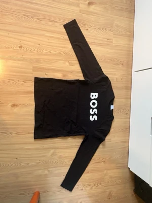 Svart långärmad BOSS t-shirt - Snygg svart långärmad t-shirt från BOSS med stor vit logga tryckt vertikalt på framsidan. Tillverkad i mjuk bomull som känns skön mot huden. Perfekt för dig som gillar stilrena och klassiska plagg med en tydlig logga.