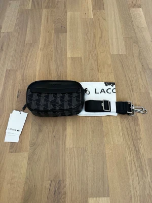 Svart axelväska från Lacoste - Snygg svart axelväska från Lacoste med geometriskt mönster och justerbar bred axelrem. Väskan har silverfärgade detaljer och dragkedja samt en liten ficka framtill. Perfekt för att bära det viktigaste och ger en stilren look.