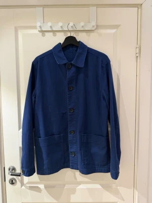 Mörkblå overshirt med stora fickor - Stilren mörkblå overshirt i bomull med klassisk krage och stora framfickor. Jackan har raka ärmar, knäppning framtill med fem knappar och enkel, minimalistisk design. Perfekt lager på lager-plagg för en avslappnad look.