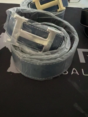 H bälte vändbart blå-svart - Snyggt svart bälte i läder med ett stort, silverfärgat H-format metallspänne. Bältet har vita kontrastsömmar längs kanterna som ger en cool detalj. Perfekt accessoar för att lyfta din outfit.