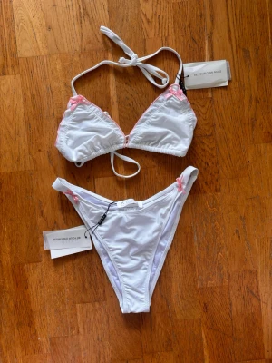 Vit bikini med rosa rosetter - Söt vit bikini med trekantsöverdel och smala axelband som knyts i nacken och ryggen. Både överdel och underdel har små rosa rosetter i hörnen. Bikinin har en enkel och stilren design med ljusrosa detaljer. Perfekt för lata dagar på stranden. Helt ny med prislapp kvar från Kaiia. Nypris 550!