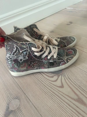Odd Molly blommiga höga sneakers - Unika höga sneakers från Odd Molly med färgglatt blommönster i grönt, rött och beige. Skorna har råa kanter, vita sulor och beige snörning. Tillverkade i tyg med broderade detaljer och logga på sidan. Perfekta för dig som gillar boho-stil.