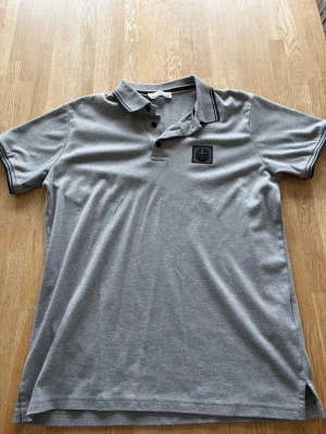 Grå stone Island pikétröja med svart logga - Klassisk grå pikétopp med svart logga på bröstet och svarta detaljer på krage och ärmslut. Toppen har korta ärmar, knappar vid halsen och en normal passform. Modellen i andra bilden är 183cm lång.