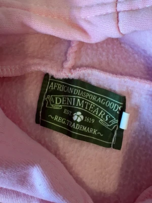 Denim tears - Säljer en oversized rosa hoodie med vita blommönster över hela tröjan. Hoodien har stor huva och en klassisk magficka framtill. Perfekt för dig som gillar färgglada och unika plagg. Materialet är mjukt och skönt, troligtvis bomullsblandning.