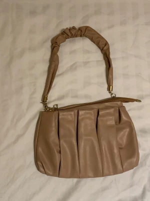 Beige axelremsväska med veckad design - Trendig beige handväska i mjukt, veckat konstläder med bred, scrunchie-inspirerad axelrem och guldfärgade metalldetaljer. Väskan har dragkedja upptill och en rymlig insida, 