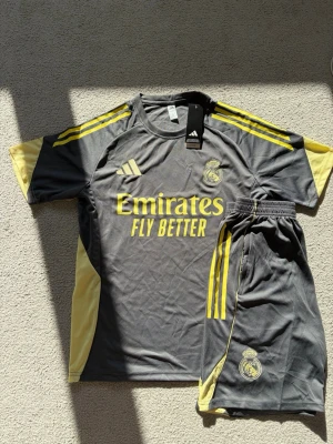 Real Madrid träningsset grå/gul Adidas - Snyggt träningsset från Real Madrid i grått med gula detaljer. Setet består av en t-shirt och shorts, båda med klubbens logga och Adidas ränder. Materialet är lätt och andas, perfekt för träning eller match. Emirates Fly Better tryck på bröstet.