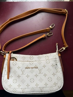 Vit axelväska  - Snygg vit axelväska med klassiskt monogrammönster i beige. Väskan har justerbar och avtagbar rem i ljusbrunt läder samt guldfärgade metalldetaljer. Perfekt för dig som vill ha en trendig och lyxig accessoar till din outfit.