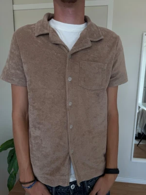 Beige frottéskjorta med kort ärm - Säljer en beige skjorta i mjuk frotté med korta ärmar och klassisk krage. Skjortan har knappar framtill och en bröstficka. Perfekt för en avslappnad och trendig look.