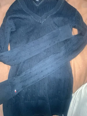 Mörkblå stickad tröja Tommy Hilfiger - Snygg mörkblå stickad tröja från Tommy Hilfiger i 100% pima bomull. Tröjan har v-ringning, diskret kabelstickat mönster och långa ärmar. Liten logga på ärmslutet ger en stilren touch. Perfekt för dig som gillar klassisk och enkel stil.