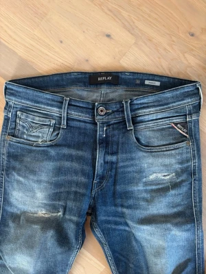 Blå slitna jeans från Replay - Säljer ett par blå jeans från Replay med slitningar och tvättade detaljer. Klassisk femficksmodell med Replay-logga på myntfickan och läderpatch bak i midjan. Jeansen har en avslappnad passform och är tillverkade i denim. Mått: midja 44, innerben 72, ytterben 92. 