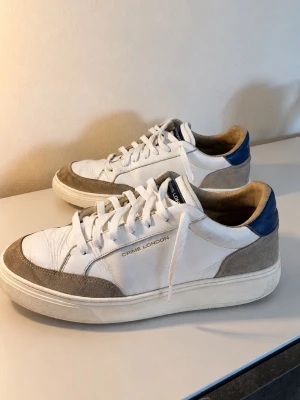 Vita sneakers från Crime London - Snygga vita sneakers från Crime London med grå mockadetaljer och blå häl. Skorna har en chunky sula och klassisk låg profil. Ovandelen är i läder och mocka, med vita skosnören och diskret logga på sidan.