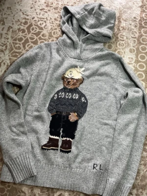 Ralph Lauren - Säljer en grå stickad hoodie med huva och ett stort broderat björnmotiv på framsidan. Tröjan har ribbade muddar och RL-broderi nedtill. Perfekt för dig som gillar unika och mysiga plagg med cool design. Tag där vid halsen gått upp några stygn
