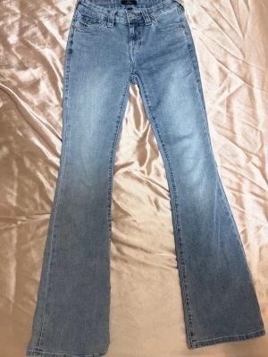 Bootcut jeans i ljusblå denim - Säljer ett par ljusblå bootcut jeans från True Religion med klassiska kontrastsömmar och detaljerade bakfickor med knappar. Jeansen har normal midja och fem fickor. Perfekta för dig som gillar en avslappnad men snygg stil.