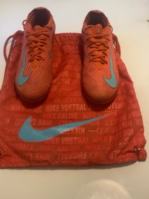 Nike vapor 16 elite mad energy pack - Säljer ett par grymma orangea Nike fotbollsskor med ljusblå swoosh på ovansidan. Skorna har snörning och är tillverkade i ett lätt syntetmaterial för bra bollkontroll. Medföljer matchande orange gympåse med Nike-logga. Perfekta för dig som vill sticka ut på planen!