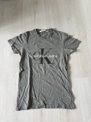 Grå Calvin Klein Jeans t-shirt - Snygg grå t-shirt från Calvin Klein Jeans med stort CK-logotyptryck på bröstet. Klassisk rund halsringning och korta ärmar. Tillverkad i mjukt bomullsmaterial som känns skönt mot huden. Perfekt för en avslappnad och trendig look.
