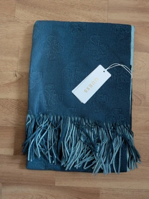 Blå sjal från Guess med fransar - Ladda upp inför hösten med denna sjal. Snygg blå sjal från Guess med diskret logomönster över hela tyget och långa fransar i ändarna. Sjalen är rektangulär och har en mjuk, lyxig känsla som passar perfekt till jackan eller kappan. Materialet känns lent och följsamt.