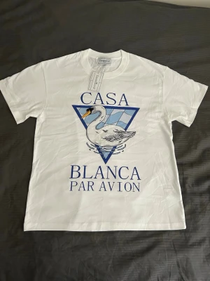 Vit Casa Blanca t-shirt med svanmotiv - Vit t-shirt från Casa Blanca med ett stort tryck av en svan och blå triangulär bakgrund på bröstet. Texten 'CASA BLANCA PAR AVION' i blått framtill. Klassisk passform och rund hals. Perfekt för dig som gillar grafiska prints.