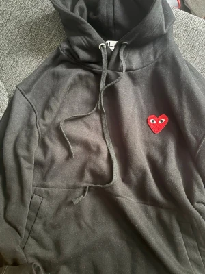 Svart comme des garcons hoodie  - Säljer en svart hoodie med huva och dragsnören. På bröstet finns ett broderat rött hjärta med ögon, vilket ger en unik och trendig look. Hoodien har magficka och är tillverkad i mjukt material, perfekt för en avslappnad stil.
