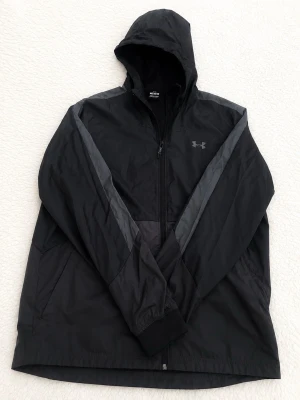 Svart vindjacka från Under Armour - Snygg svart vindjacka från Under Armour med huva och dragkedja framtill. Jackan har grå detaljer längs ärmarna och meshfoder på insidan för extra komfort. Perfekt för blåsiga dagar och sportiga outfits.