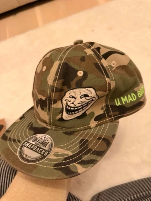 Camouflage keps med trollface och text - Säljer en snapback-keps i camouflage med broderad trollface-meme på framsidan och texten 'U MAD BRO?' på sidan. Kepsen har klassisk böjd skärm och justerbar snapback. Perfekt för dig som gillar memes och streetstyle.