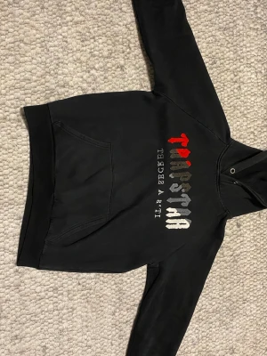 Svart Trapstar hoodie med tryck - Säljer en svart hoodie från Trapstar med färgglatt broderat tryck på bröstet i rött, grått och vitt. Klassisk känguruficka framtill och huva med dragsko. Perfekt för dig som gillar streetwear och vill ha en skön och stilren hoodie.